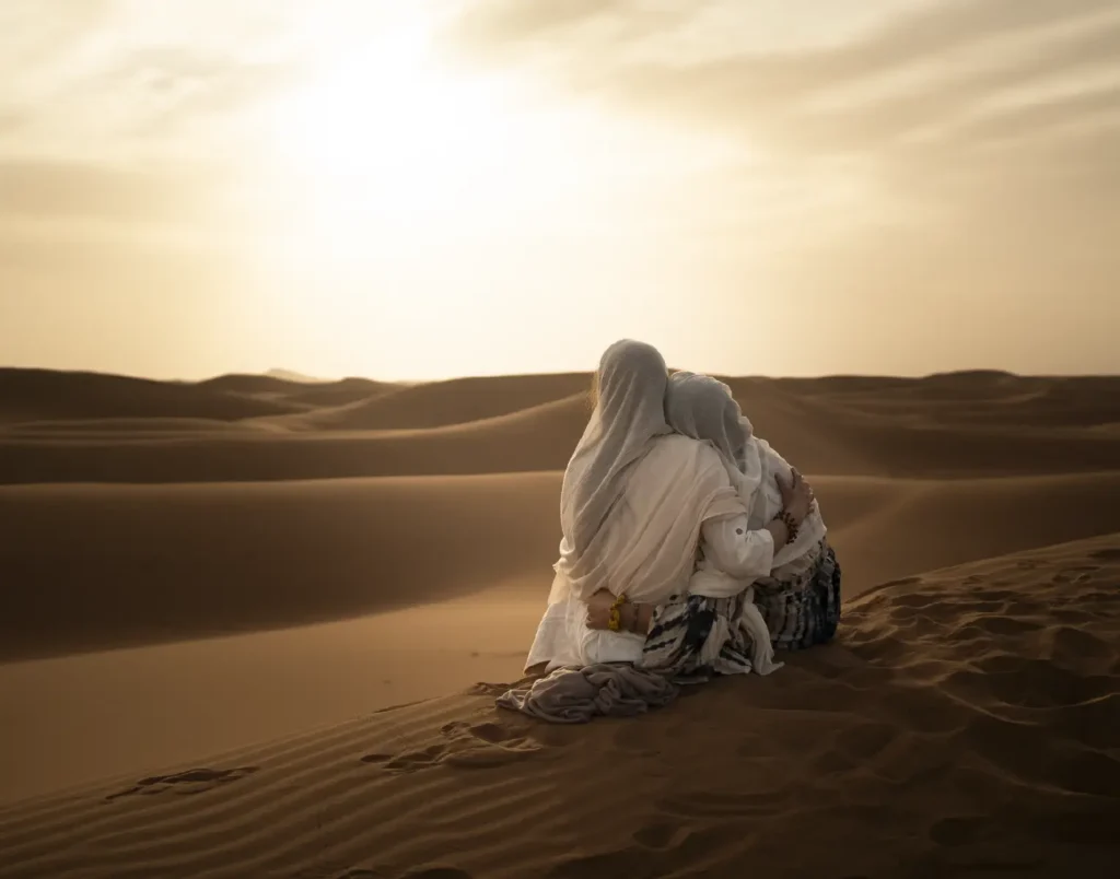 Due persone abbracciate, sedute sulle dune nella luce del sole.
