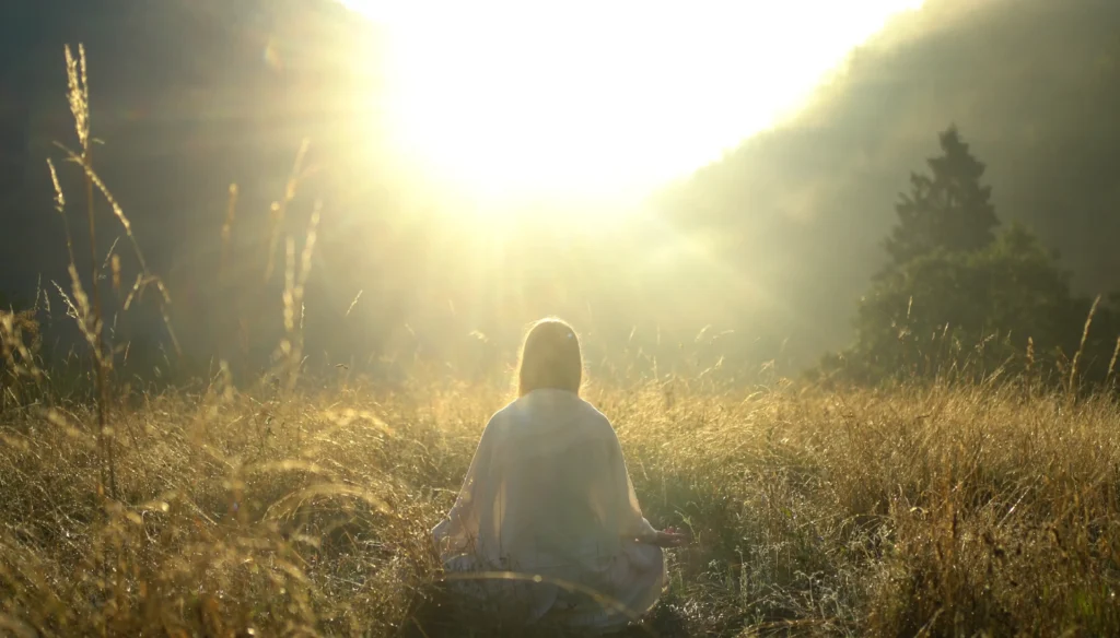 Giulia seduta di spalle in meditazione in un prato illuminato dal sole.