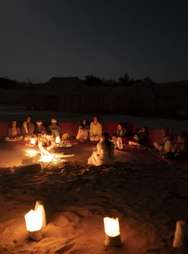 Un gruppo seduto in cerchio nel deserto, intorno al fuoco e alle candele, di notte.
