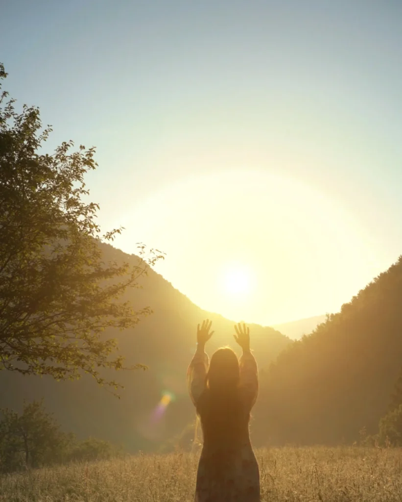 Giulia in piedi nel prato con le braccia alzate verso il sole, tra le montagne.