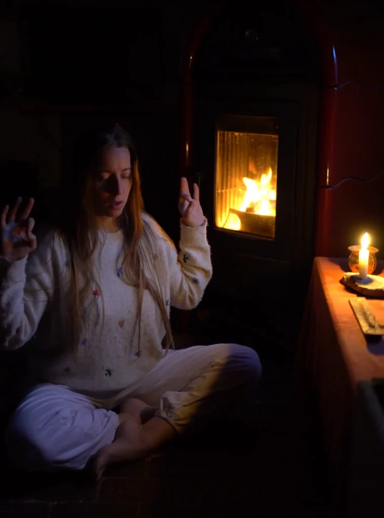 Giulia seduta a occhi chiusi accanto al fuoco, con le mani sollevate.